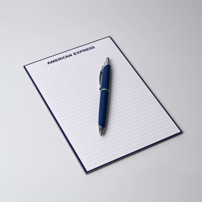 A5 notepad with 50 lined leaves, featuring a blue cover and a silver pen placed on top. It has a logo at the top.