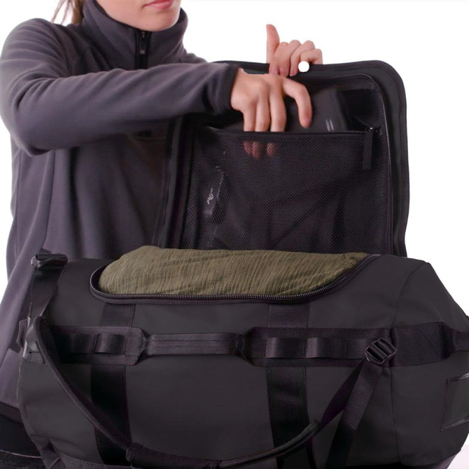 A black duffle bag with a mesh pocket and a rolled green item inside, being held by a person in a grey jacket.
