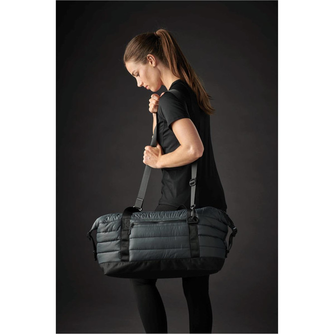 A grey quilted duffle bag with adjustable straps, being carried by a person in athletic wear.