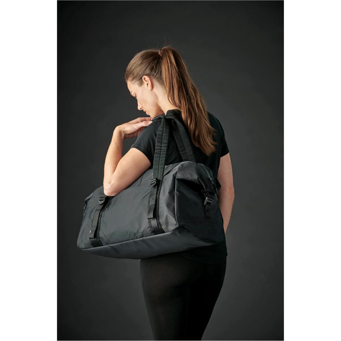 A woman carrying a large black duffle bag over her shoulder. The bag has a logo and adjustable straps.