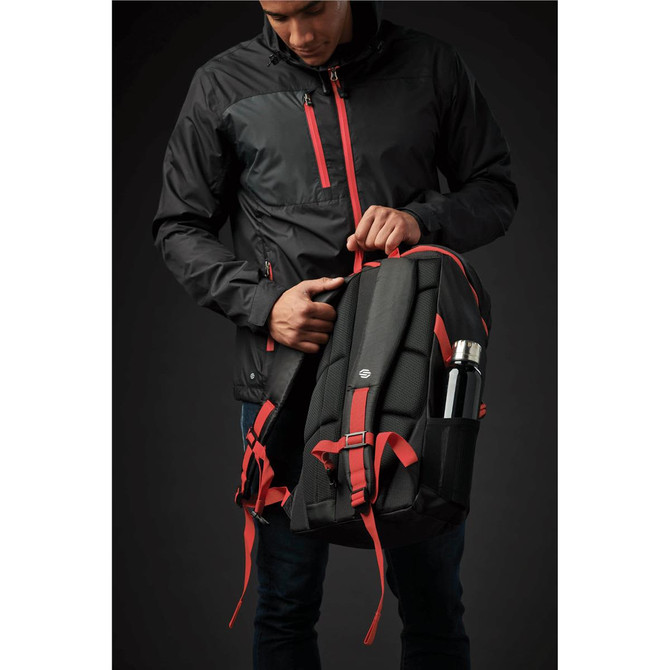 A black and red backpack with padding, side pockets, and a drink bottle holder, held by a person in a jacket.