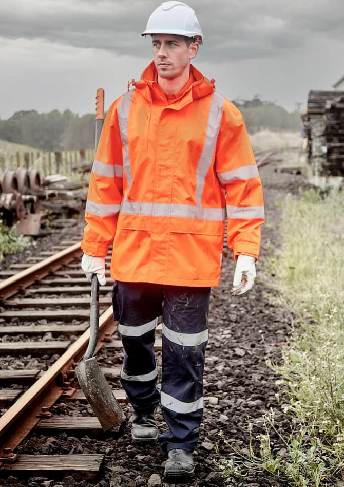 A man wearing an orange waterproof jacket and navy trousers walks along railway tracks, holding a tool. The jacket features reflective stripes.