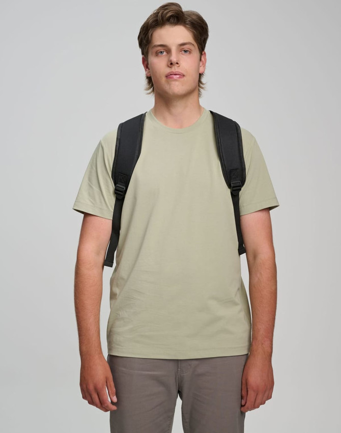 A man wearing a light khaki T-shirt and grey pants, carrying a black backpack with shoulder straps.