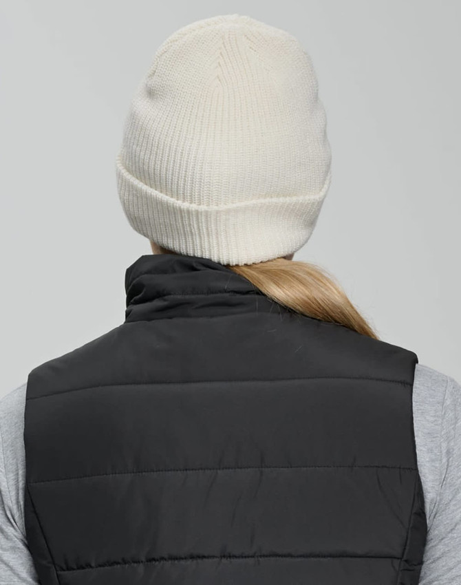 A cream beanie with a ribbed design, worn by a person in a black vest. The back of the beanie is visible.