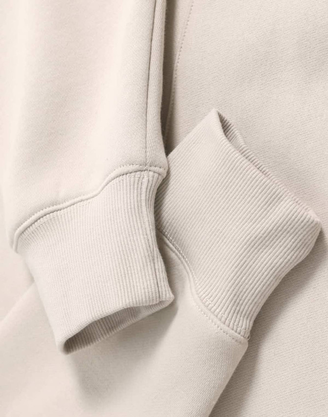 A close-up of a beige oversized hoodie featuring ribbed cuffs and a soft fleece texture. It has a logo.