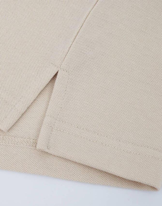 A close-up of a beige cotton/poly pique polo shirt, highlighting the stitched hem and sleeve detail.