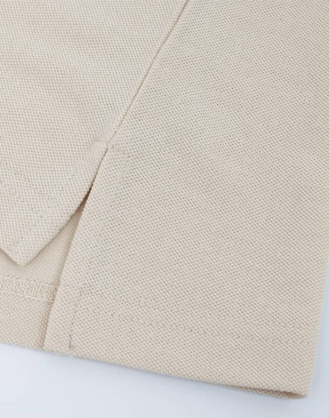 A close-up of a beige cotton/poly pique fabric, showcasing its texture and stitching details. It features a simple hemline.