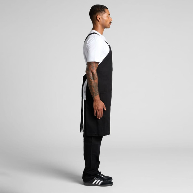 A black apron is worn by a person standing sideways, featuring adjustable straps and a logo.