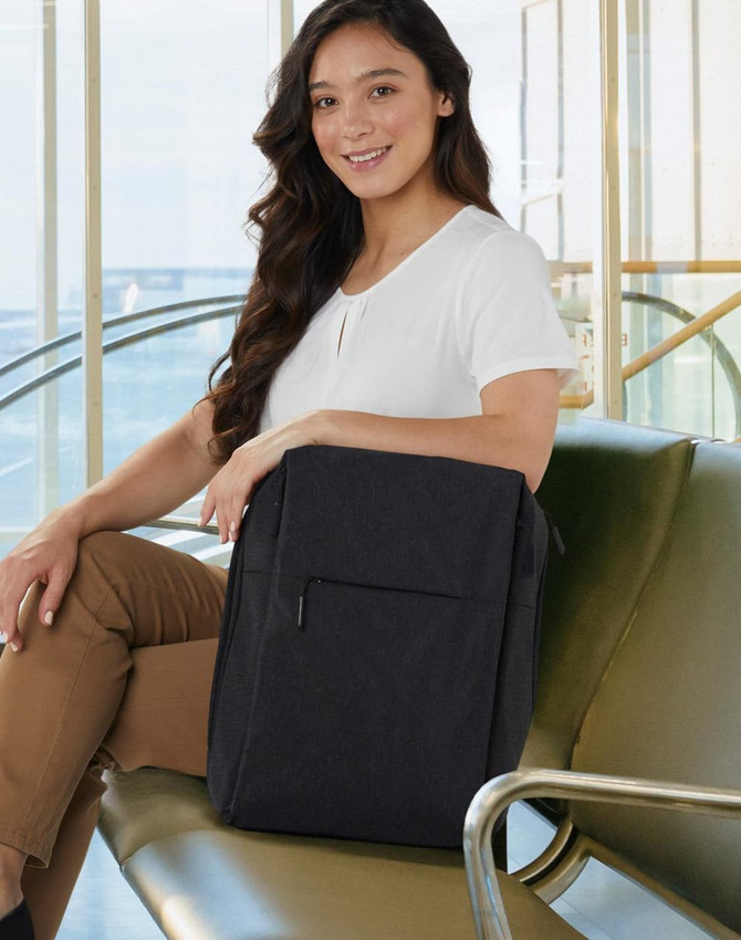 A black backpack with a front pocket, shown on a woman sitting on a green bench in a bright, modern environment.
