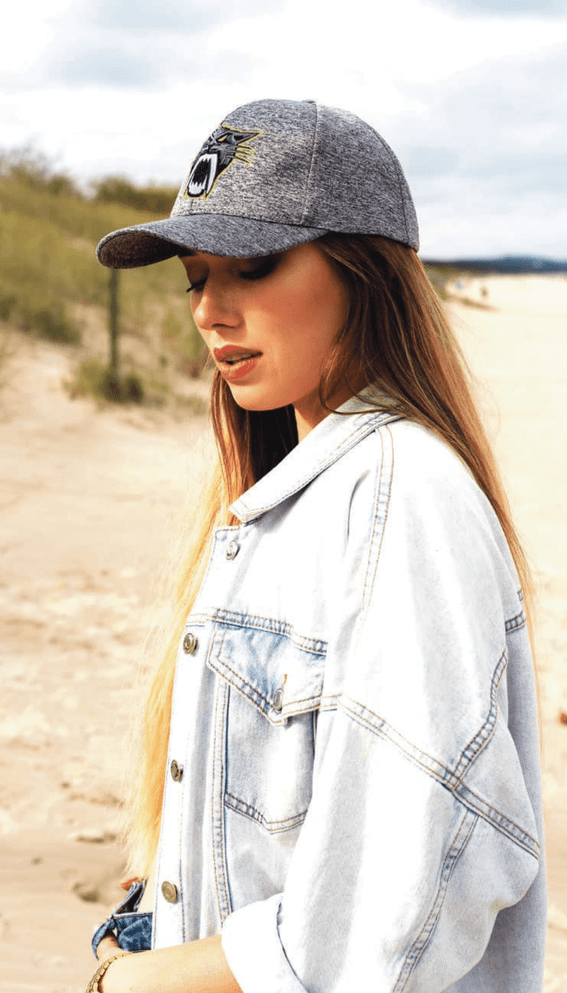 A woman wearing a grey cap and a denim jacket stands on a sandy beach, with dunes in the background.