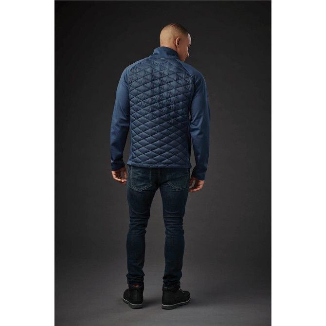 A man wears a navy blue thermal shell jacket with a quilted back, paired with dark jeans. The jacket has a logo.