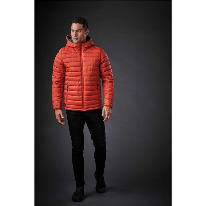 A man wearing an orange thermal jacket with a hood, paired with black trousers and shoes, against a dark background.