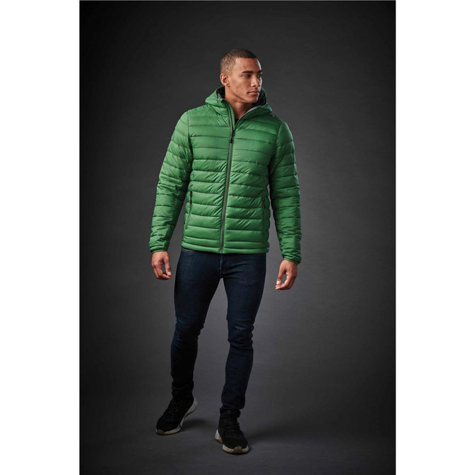 A man wearing a green thermal jacket with a hood, paired with dark jeans and black shoes. The jacket has a logo.