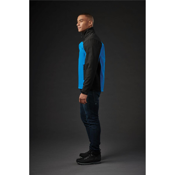 A man wearing a blue and black softshell jacket, standing sideways against a dark background.