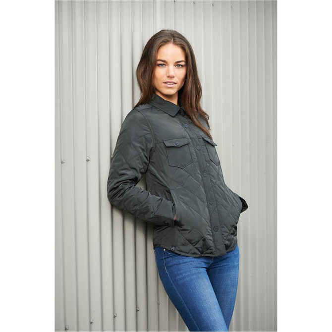 A woman leans against a wall wearing a dark green quilted jacket with buttons and chest pockets.