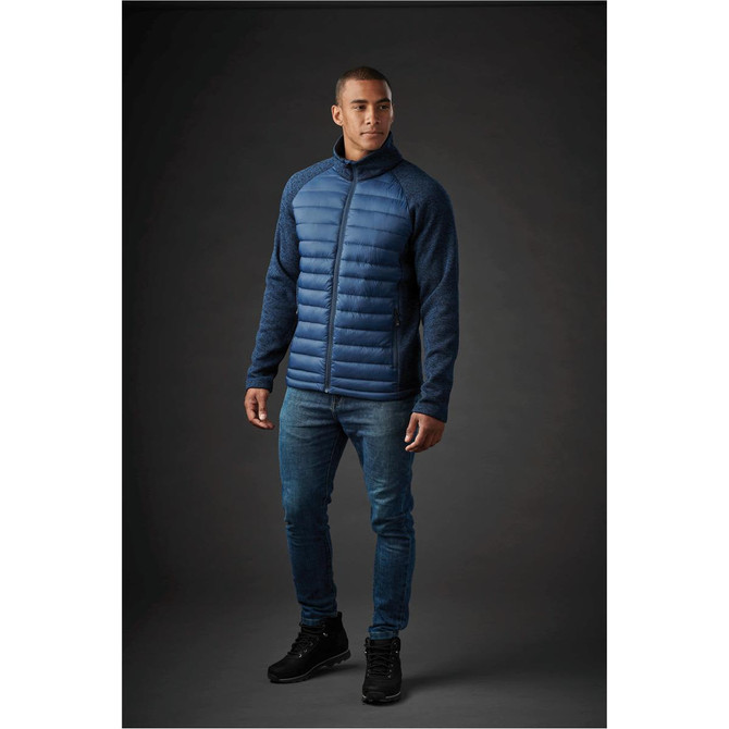 A man wearing a blue hybrid jacket with a padded front and textured sleeves, paired with jeans and boots.