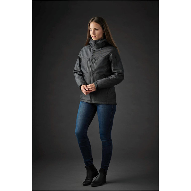 A woman stands in a black Women's Epsilon System Jacket, paired with blue jeans, against a dark background. The jacket features a logo.