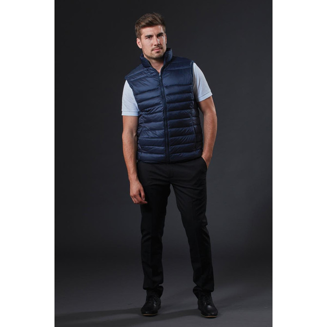 A man wearing a navy puffer vest over a white shirt, paired with black trousers and shoes, standing against a dark background.