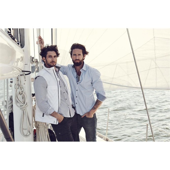 Two men posing on a sailboat, one wearing a light blue button-up shirt and the other in a white vest over a checked shirt.