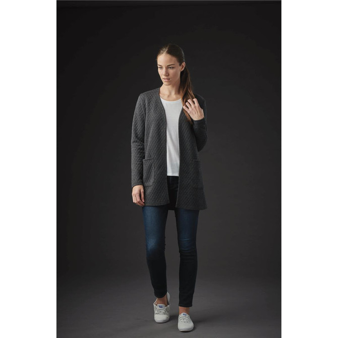 A woman wearing a grey quilted cardigan over a white top and jeans stands against a dark background.
