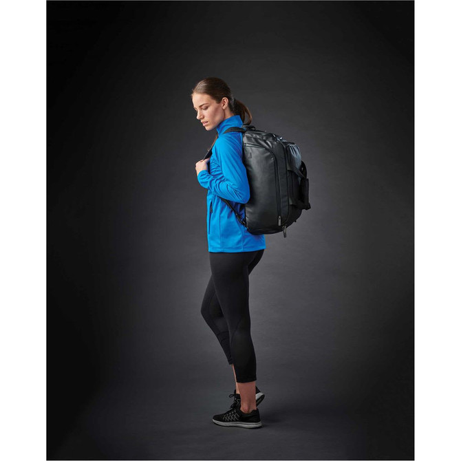 A woman in a blue lightweight softshell jacket, wearing black leggings and a backpack, standing against a dark background.