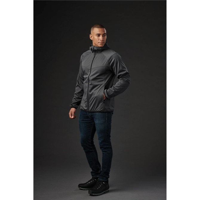 A model wearing a grey men's softshell jacket, paired with dark jeans and black shoes, standing against a dark background.
