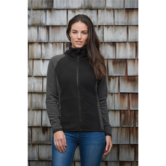 A woman wears a black and grey zip-up microfleece jumper, posing against a wooden background. The jumper features a logo.