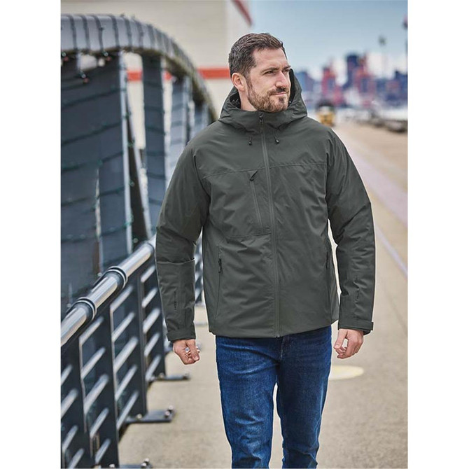 A man wearing a dark green thermal shell jacket, walking outdoors along a paved path. The jacket features a hood.