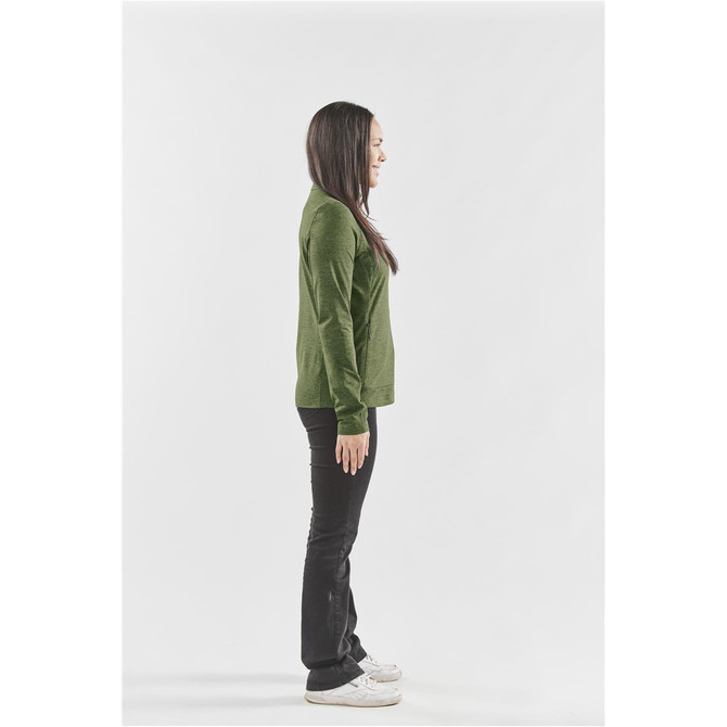 A woman stands sideways wearing a green performance jacket and black pants, with white shoes. The jacket has a logo.