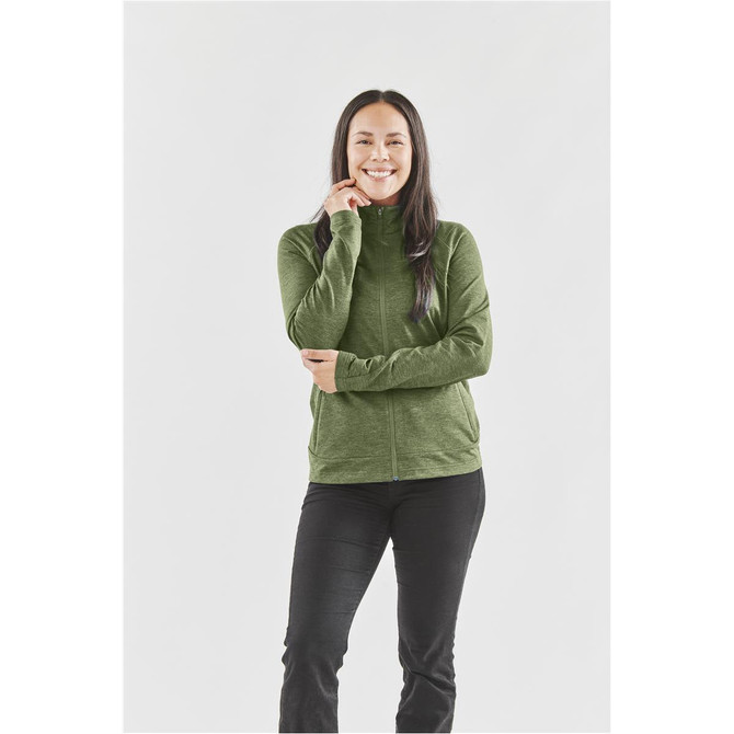 A woman stands smiling in a green performance jacket, paired with black pants. The jacket features a high collar and a logo.