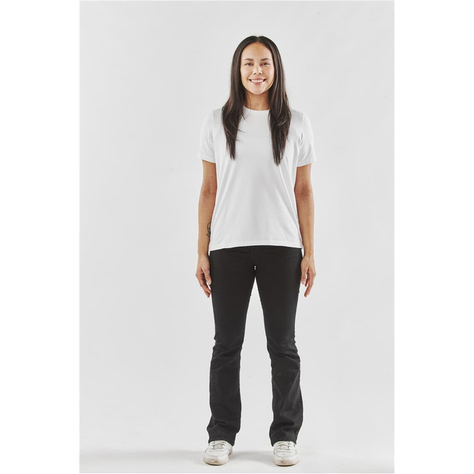 A woman stands in a fitted white t-shirt and black pants, with a plain background. The shirt has a logo.