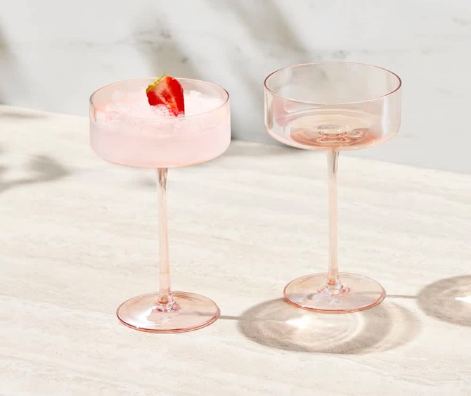 A set of two pink coupe glasses, one filled with a drink and garnished with a strawberry, displayed on a light surface.