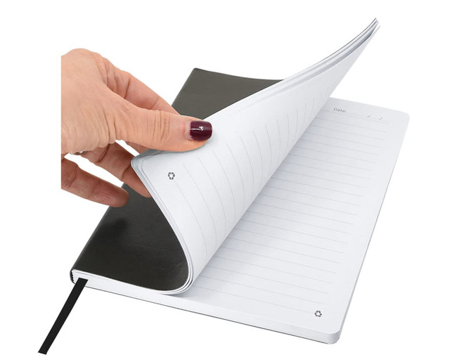 A hand holding an A5 notebook with a black cover, showing blank lined pages inside. The notebook has a ribbon bookmark.