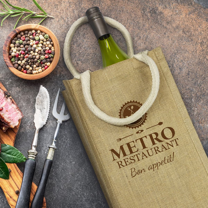 A jute double wine carrier in beige, with a bottle inside, accompanied by utensils and a bowl of mixed peppercorns.