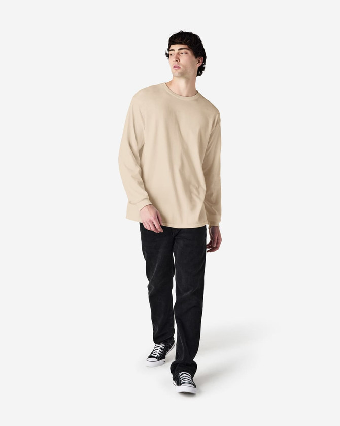 A man is wearing a beige long sleeve T-shirt paired with black jeans and sneakers, featuring a logo.