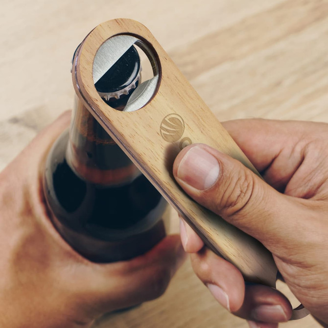 A drink bottle opener made of wood and metal, being used to open a dark beverage bottle. It features a logo.