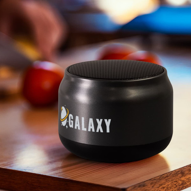 Auris Bluetooth speaker in black, featuring a textured top and a visible logo, placed on a wooden surface near apples.