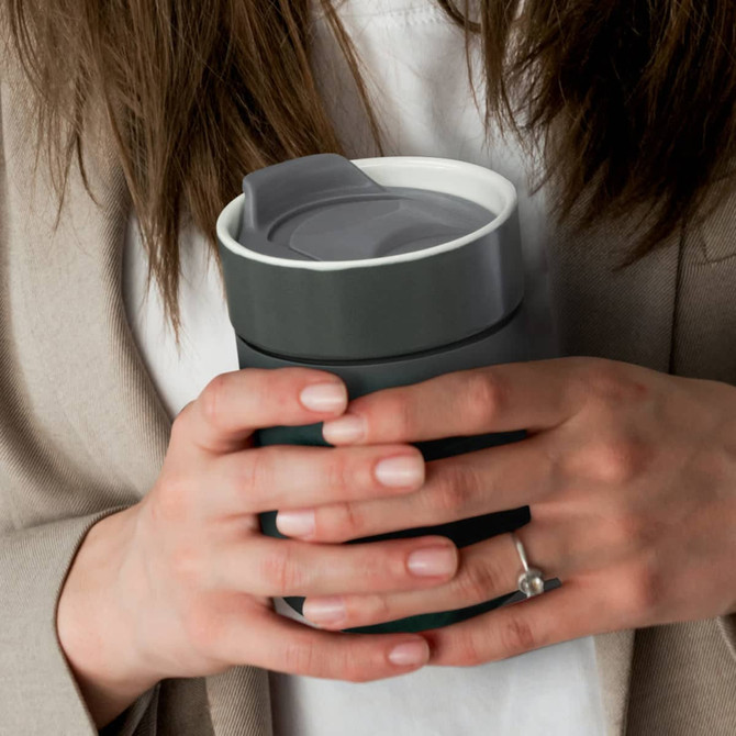 A ceramic coffee cup in dark green with a grey lid, held in two hands. Features a logo on the side.