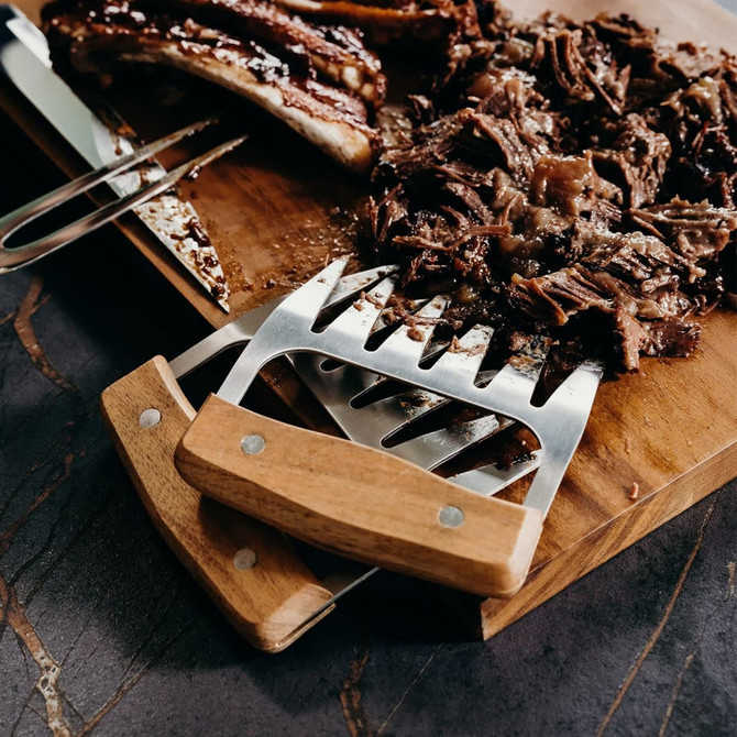 Keepsake Meat Shredding Claws with stainless steel prongs and a wooden handle, resting on a cutting board beside pulled meat.
