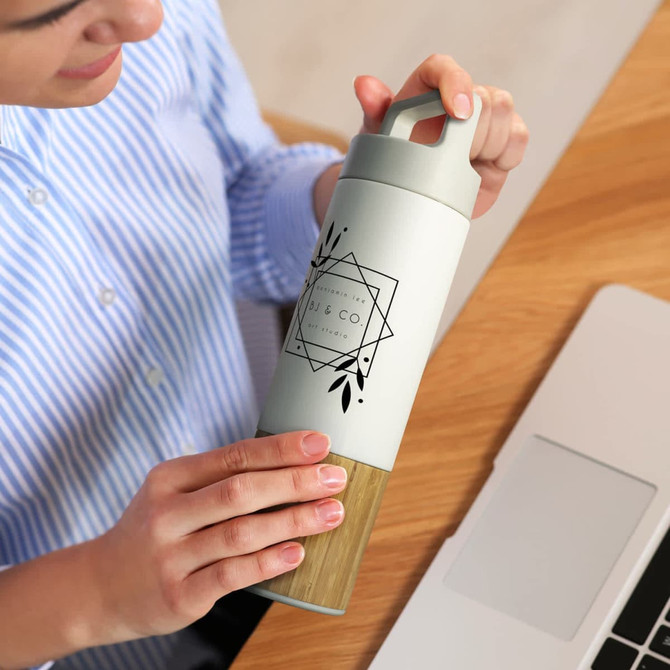 A drink bottle in green and wood finish features a logo and a secure handle, held by a person near a laptop.