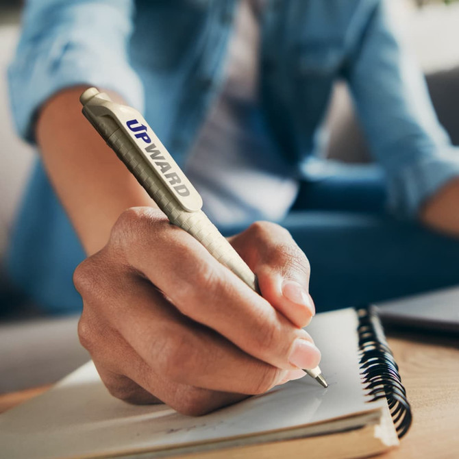 A grey and silver pen is being held in a person's hand, poised to write in a notebook. The pen has a logo.