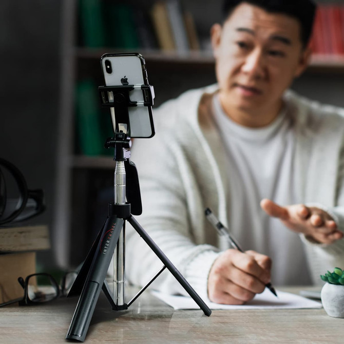 A black selfie stick tripod holds a smartphone, with a person writing in the background. The setting is casual.