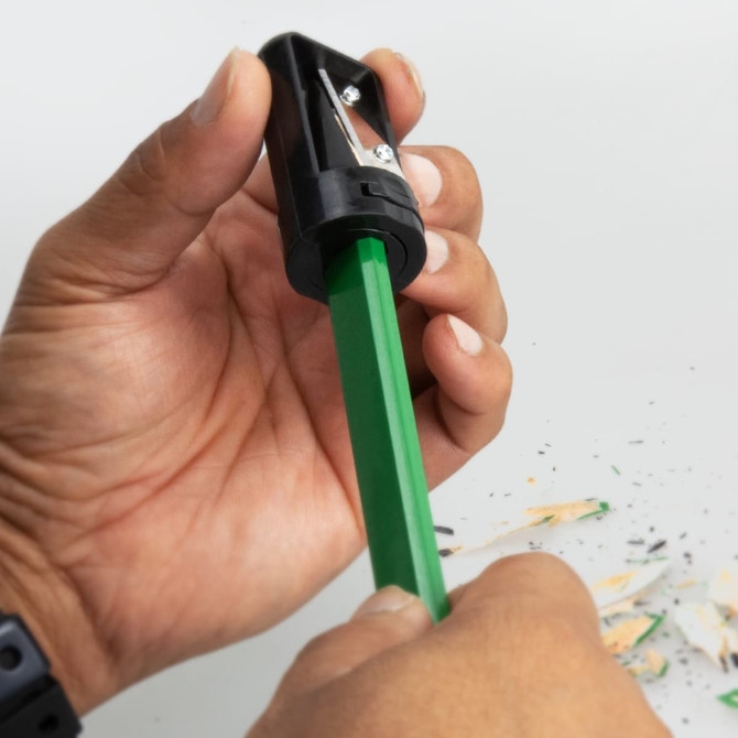 A black and grey pencil sharpener being used on a green carpenter’s pencil, surrounded by wood shavings.