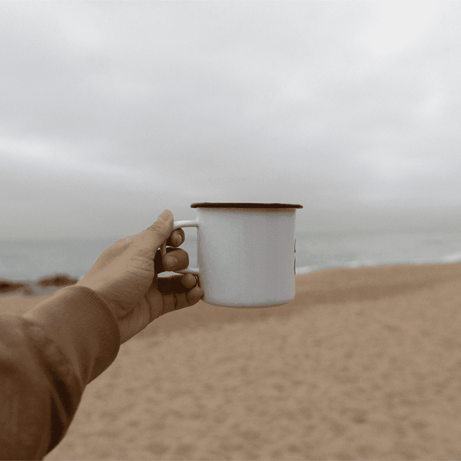 A drink bottle featuring a white enamel metal cup with a black rim, held against a beach backdrop.