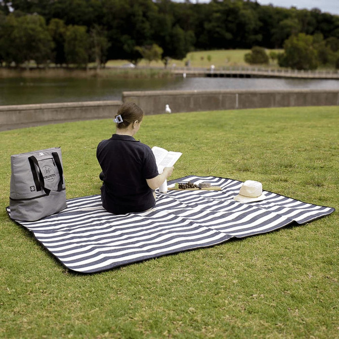 A striped navy and white picnic rug with waterproof backing, laid out on grass, with a person reading on it.