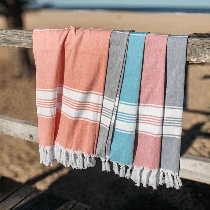 A set of four Turkish towels in coral, blue, grey, and pink, featuring fringed edges and laid over a wooden railing.