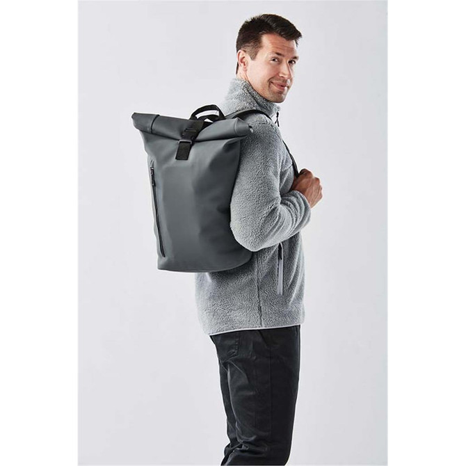A man wearing a grey fleece jacket carries a dark grey backpack with a roll-top design. The backpack has a logo.