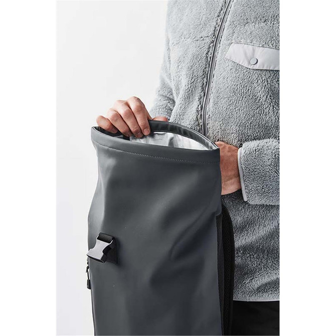 A person holds a roll-top backpack in dark green, featuring a silver interior and a logo on the side.