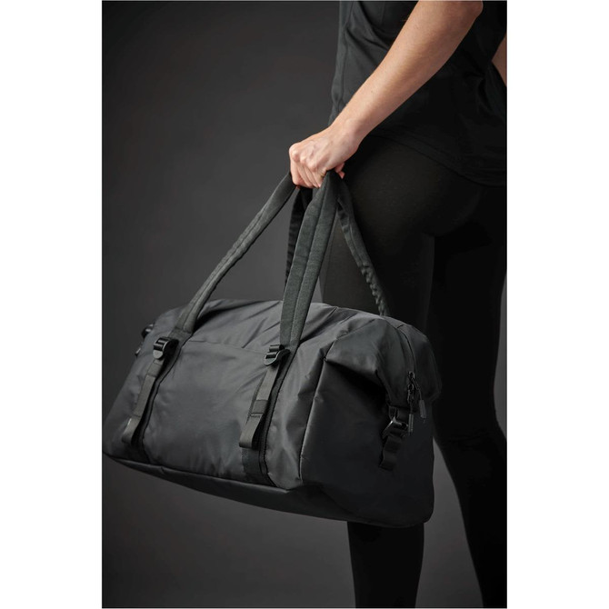 A person holds a black duffle bag with braided handles, featuring buckles and a sleek design.