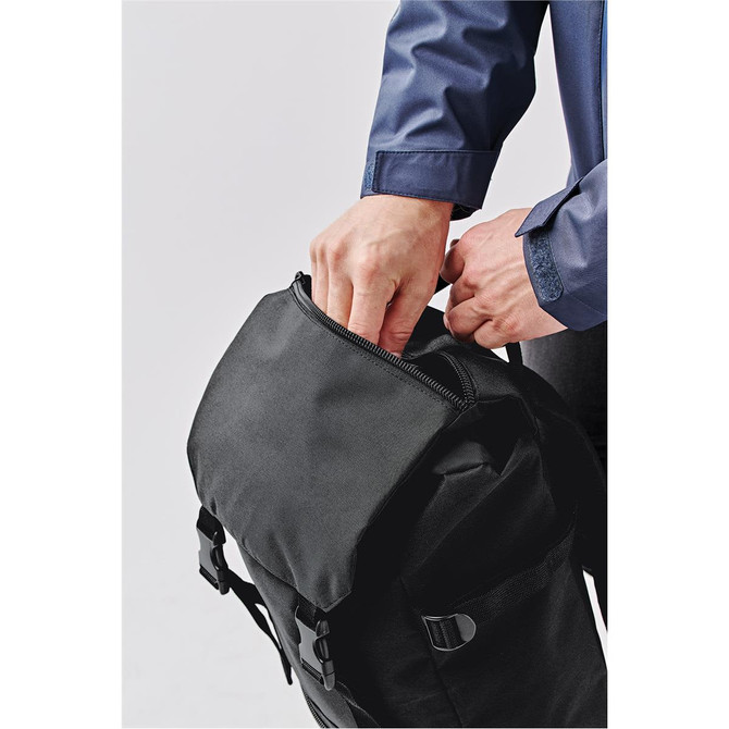 A black backpack with a zippered opening, being handled by a person in a blue shirt. It features adjustable straps and buckles.